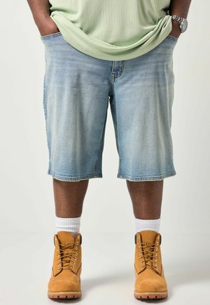 Person iført lyse denimshorts, hvide sokker, lysebrune arbejdstøvler og en lysegrøn skjorte med hænderne i lommerne.