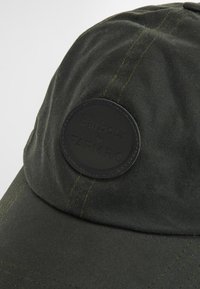 Dark green cap made of a textured fabric with visible stitching and a circular black leather patch featuring the Barbour and FARM RIO logos.