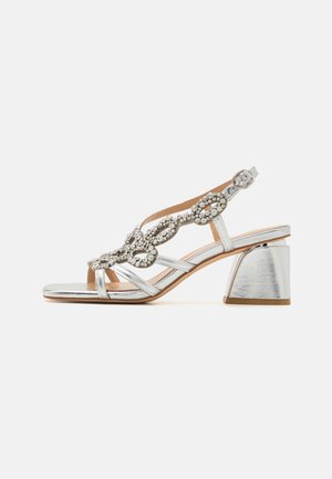 Sandalias plateadas con tiras, que cuentan con un bajo tacón cuadrado, adornadas con un diseño de joyas en la parte superior y una correa ajustable en el tobillo.