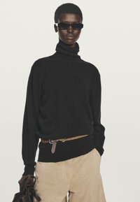 Person, der einen schwarzen Rollkragenpullover, beige Hose mit einem braunen Gürtel, schwarze Sonnenbrille trägt und eine dunkelbraune Ledertasche hält.