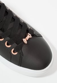 Černé kožené tenisky s černými tkaničkami, růžově zlatými kovovými očky a mašlí. Bílá gumová podrážka s hladkým povrchem.