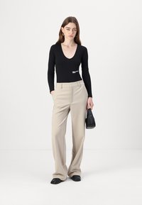 Black long-sleeve bodysuit with a v-neck and logo detail, paired with beige wide-leg trousers. Black handbag and simple black shoes.
