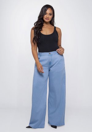 Vrouw met lang donker haar, gekleed in een zwart tanktop en lichtblauwe wijde jeans, staand tegen een effen witte achtergrond.