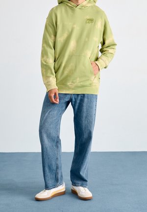 Osoba ubrana w lime'ową bluzę z kapturem typu tie-dye z logo Puma, niebieskie dżinsy i białe trampki stoi na niebieskiej podłodze na tle białej ściany.