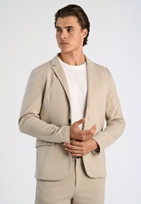 Blazer beige su misura in tessuto morbido con chiusura a un bottone, taschino frontale e pantaloni abbinati, indossato sopra una camicia bianca.