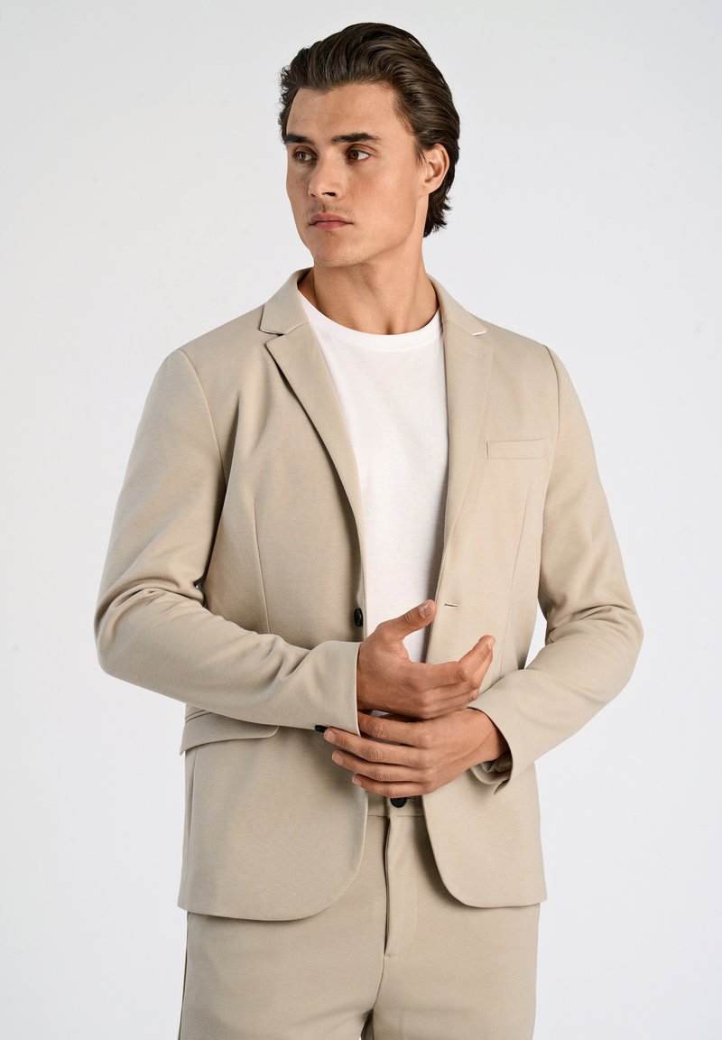Blazer beige su misura in tessuto morbido con chiusura a un bottone, taschino frontale e pantaloni abbinati, indossato sopra una camicia bianca.