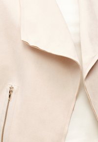 Φωτεινό μπεζ παλτό με φαρδύ γιακά και χρυσλό φερμουάρ στην αριστερή πλευρά, εμφανιζόμενο σε κοντινή λήψη λεπτομέρειας.