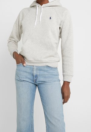 Person wearing a light gray hoodie with a small logo and light blue denim jeans, one hand in pocket, against a plain background.