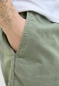 Pantalones cortos de tela verde claro con una costura de bolsillo visible. El material parece suave, con una ligera textura. Primer plano de la mano descansando en el bolsillo.