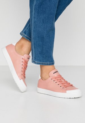 Osoba w skróconych niebieskich jeansach i jasnoróżowych skórzanych sneakersach z białymi podeszwami, stojąca na jednolitym jasnym tle.