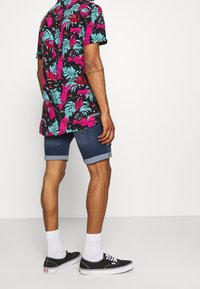 Mies, joka pukeutunut musta- ja vaaleanpunakukalliseen lyhythihaiseen paitaan, tummansiniset, lahkeet ylöskäännettyinä shortsit, valkoiset sukat ja mustat kangaskengät, seisoo sivuttain.