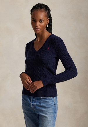 Femme aux cheveux tressés portant un pull en maille torsadée bleu marine à col en V et un jean bleu clair, debout les mains jointes devant elle.