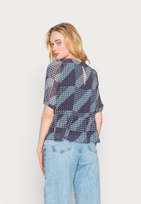 Top peplum em tecido azul marinho transparente com um padrão geométrico, apresentando mangas curtas e uma abertura em forma de olho de fechadura nas costas. Combinado com jeans azul claro.
