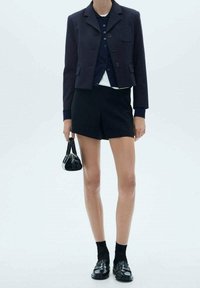 Marineblauer, taillierter Blazer über einem dunkelblauen Hemd, kombiniert mit schwarzen Shorts. Die Accessoires bestehen aus einer kleinen schwarzen Handtasche und schwarz-gelben Loafers mit Socken.