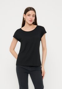 Γυναίκα με μακριά καστανά μαλλιά, φοράει χρυσούς κρίκους σκουλαρίκια, μαύρη κοντομάνικη μπλούζα και μαύρο παντελόνι, στέκεται μπροστά σε λευκό φόντο.