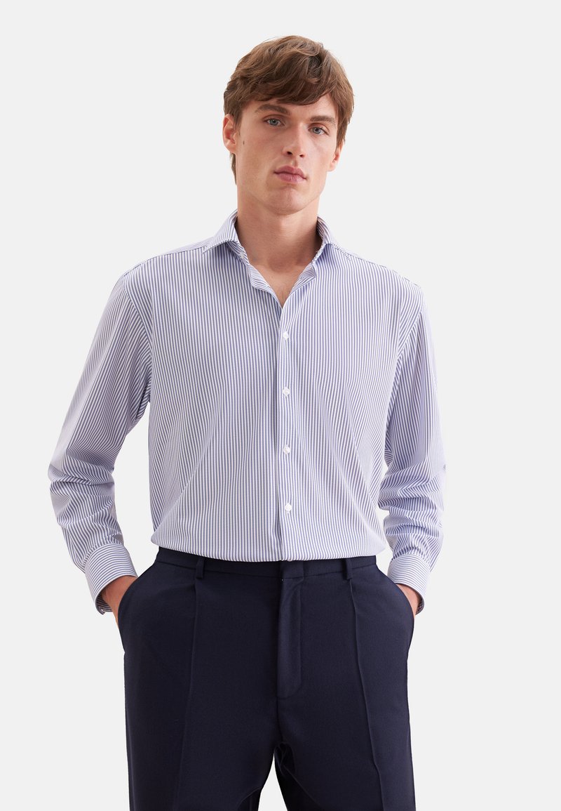 Jeune homme portant une chemise fine à rayures bleu clair et blanc avec boutons et un pantalon bleu marine foncé, debout les mains dans les poches.