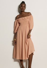 Femme portant une robe midi pêche épaules dénudées avec manches bouffantes, boucles d'oreilles anneaux dorés et bracelets dorés, debout contre un mur uni.