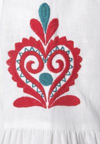 Witte stof met een rood en teal geborduurd hartontwerp, versierd met spiralen, bladaccenten en geometrische patronen. Geleefd en kleurrijke details.
