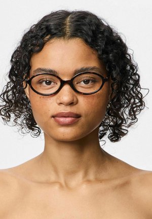 Lühikeste lokkidega naine, kes kannab ümmarguseid kilpkonnakujulisi prille, vaatab otse ette neutraalse ilmega ühevärvilise tausta ees.