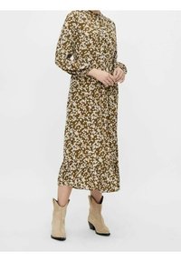 Kvinna i en brun midiklänning med vita blommönster och beige cowboystövlar med medelhögt klack, stående mot en enhetlig bakgrund.