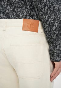 Beżowe jeansy z denimu z naszywką skórzaną na pasku, z detalicznym przeszyciem i tylną kieszenią, zestawione z wzorzystą ciemną koszulą.