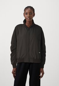 Tmavohnedá bomber bunda s vysokým golierom, zipsom a rebrovanými manžetami. Hladký, polstrovaný materiál s bočnými vreckami a logom.