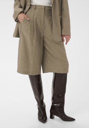 Hose: hellbrauner Stoff mit Nadelstreifen, lockere Passform, mittlere Oberschenkellänge. Stiefel: hochglänzendes, dunkelbraunes Leder mit Krokoprägung, spitze Zehen, Blockabsatz.