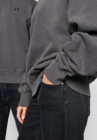 Sivý bavlnený sweatshirt s rebrovanými manžetami a malým logom na hrudi, skombinovaný s čiernymi džínsami z denimového materiálu, ktoré majú predné vrecko a detail na gombíku.