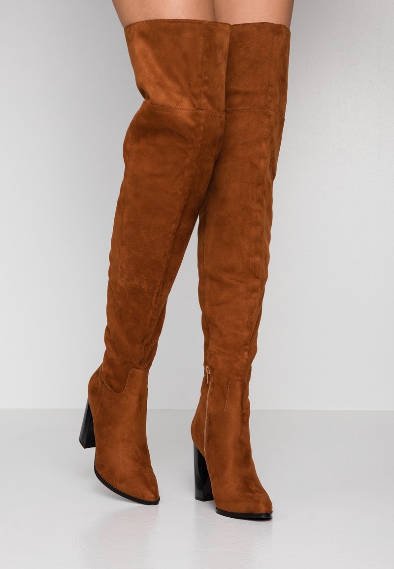 Bottes en daim brun au-dessus du genou avec un bout pointu et un talon haut noir épais. Fermeture par une fermeture éclair latérale. Texture lisse.