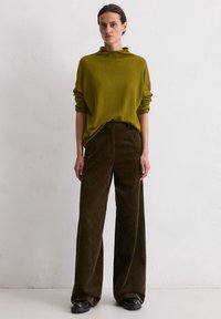 Olivgrüner Strickpullover mit hohem Kragen, kombiniert mit weiten braunen Cordhosen mit vertikalen Streifen und schwarzen Schuhen.