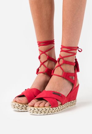 Sandales compensées rouges avec des lanières croisées autour de la cheville, en daim, présentant une plateforme tissée texturée et un petit accent métallique sur la lanière avant.