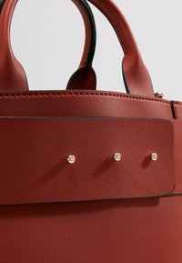 Sac à main en cuir rouge avec une forme structurée, présentant une texture lisse, des bordures noires et trois clous dorés sur un détail de sangle plate.