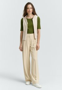 Zelený krátký rukáv, světle béžová bezrukávová vesta, široké krémové kalhoty a bílé tenisky. Hladký materiál, pohodlný outfit.