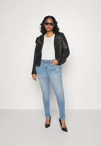 Veste en cuir noir de biker sur un t-shirt blanc, associée à un jean skinny bleu clair et des talons hauts noirs. Design simple et moderne.