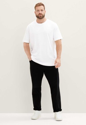 Mann mit Bart, der ein weißes Kurzarmhemd, schwarze Hose und weiße Turnschuhe trägt, steht vor einem schlichten hellen Hintergrund.