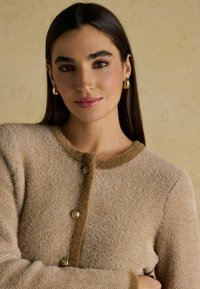 Beež kardigan, millel on kaldribade muster, pehme tekstuur, ümmargune kaelus ja kuldsed nupud; kaeluse ümber on pruun äärestus.