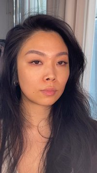 Ung kvinna med långt svart hår, neutralt ansiktsuttryck, bär ett litet halsband och ringformade örhängen, med ett fönster i bakgrunden.