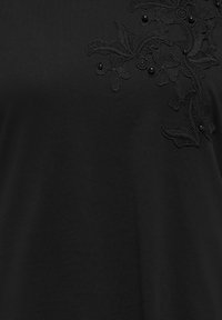 Svart stoff med blomsterapplikasjon i blonder og små runde perler på øvre høyre side.