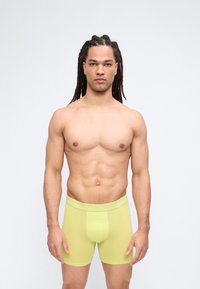 Μυώδης άνδρας με dreadlocks στέκεται με το πρόσωπο προς τα εμπρός φορώντας ανοιχτοπράσινα μποξεράκια Calvin Klein σε απλό φόντο.