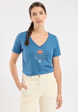 Femme aux cheveux tressés portant un t-shirt bleu col en V avec un texte en français et un motif soleil, associé à un pantalon crème taille haute.
