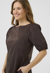 Rjava obleka s sproščenim krojem, okroglo ovratnikom, napihnjenimi rokavi na elastiko in stranskimi žepi. Narejena iz lahkotnega, gladkega materiala.