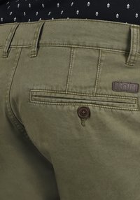 Olivengrønne bukser med en struktureret overflade, der har en baglomme med en brun knap og et læderlogo-patch på taljen.