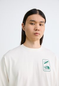 Cremefarvet langærmet t-shirt lavet af bomuld, med et grønt logo-patch på venstre brystområde. Enkelt design, rund halsudskæring.