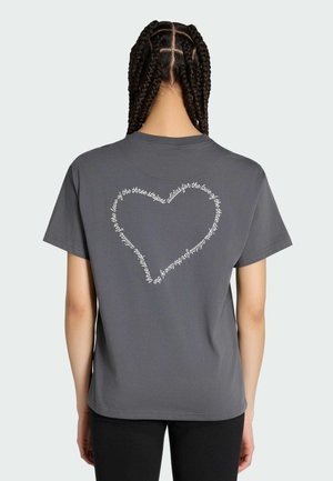 Personne aux cheveux tressés porte un t-shirt gris foncé avec un texte blanc en forme de cœur dans le dos indiquant "adidas for the love of the three stripes."