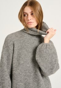 Pull en tricot gris avec un col montant, texture côtelée et manches volumineuses. Fabriqué dans un matériau doux, offrant une coupe ample.