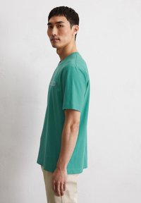 Korte mouwen, teal katoenen T-shirt met een ronde halslijn en subtiele logo-details op de borst, gedragen met lichtgekleurde pantalons.