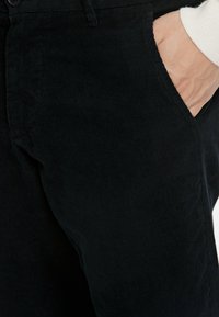 Schwarze Cordhosen mit vertikalen Rippungen, ausgestattet mit einer Vordertasche und einem Knopfverschluss. Der Stoff hat ein weiches, strukturiertes Aussehen.