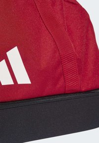 Crvena sportska torba s bijelim logotipom, otpornom tkaninom, teksturiranom površinom i crnim patentnim zatvaračem na donjem dijelu.