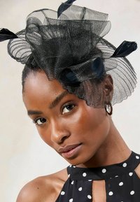 Crni mrežasti fascinator s složenim peteljkama i perjem, nošen uz top s halter izrezom s točkicama. Prikaz u blizini koji naglašava teksturu i detalje.