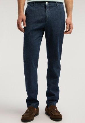 Mann trägt dunkelblaue Slim-Fit-Jeans und braune Wildleder-Loafers, steht mit entspannten Händen seitlich vor einfachem Hintergrund.